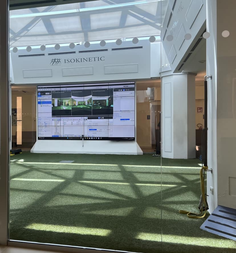 The motion-capture hall at Isokinetic Bologna: indoor turf under a glazed roof, the clinic name above the entrance, and a wall of screens running gait-analysis software