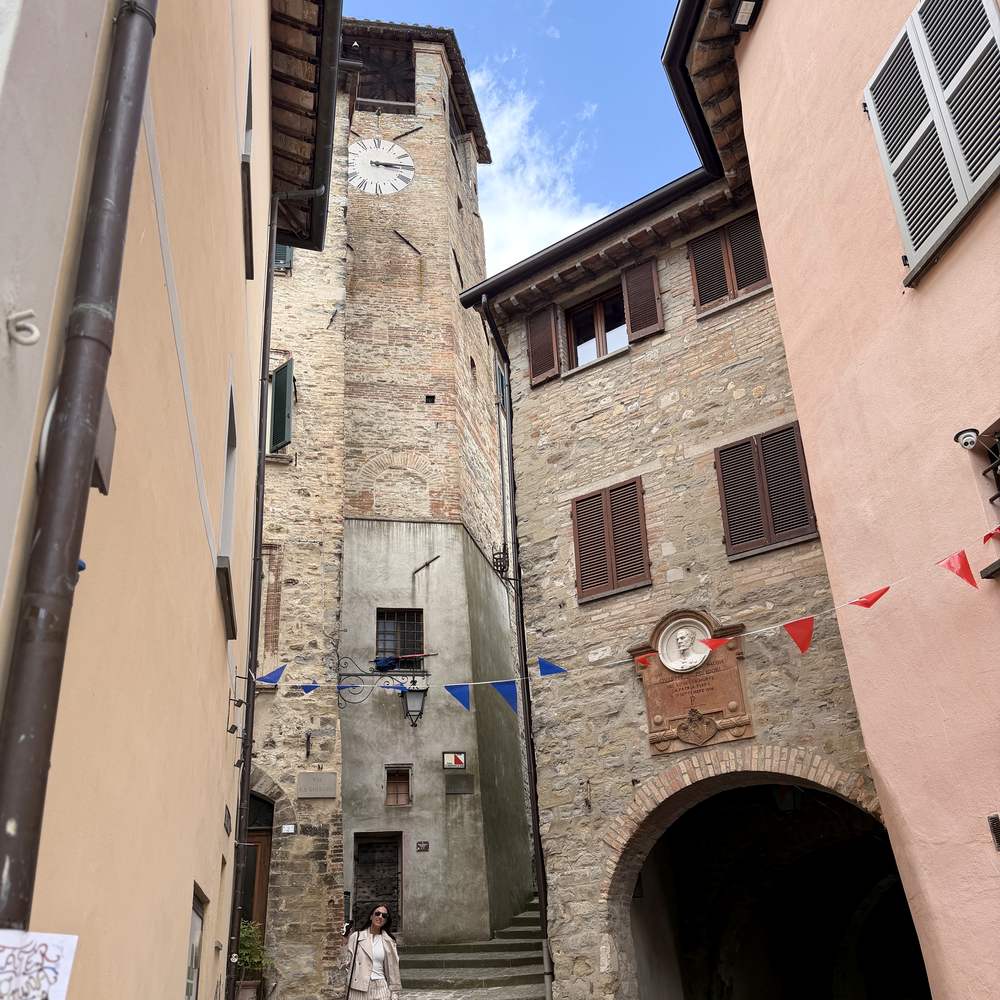 A narrow street in an Umbrian hill town, a medieval clock tower above a stone archway with bunting strung between the buildings