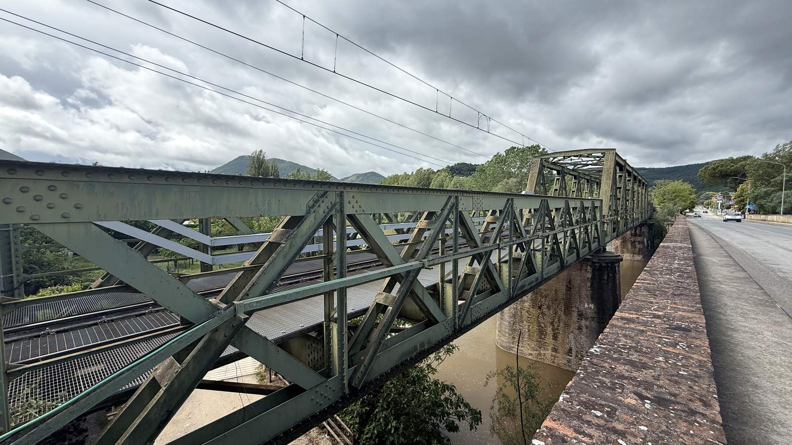 A riveted iron railway bridge carrying the line across a river, wooded hills rising behind it under a heavy sky